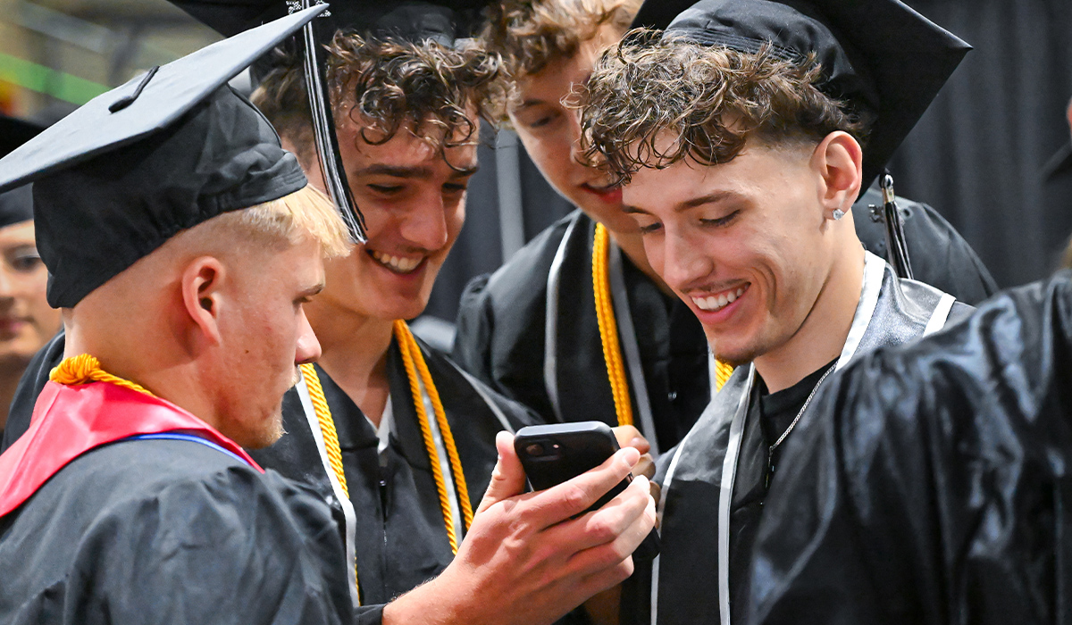 Several graduates wearing caps and gowns look down at a phone screen.