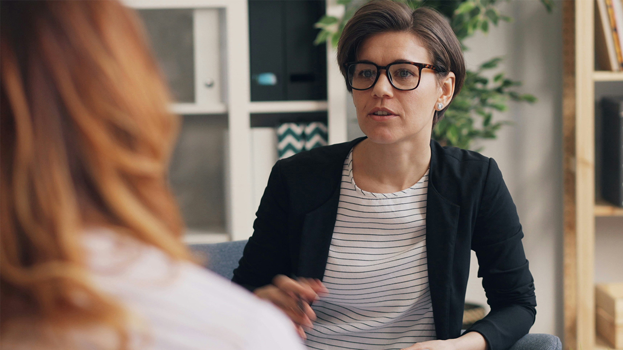 A therapist sitting on a couch talks with a patient sitting across from her.