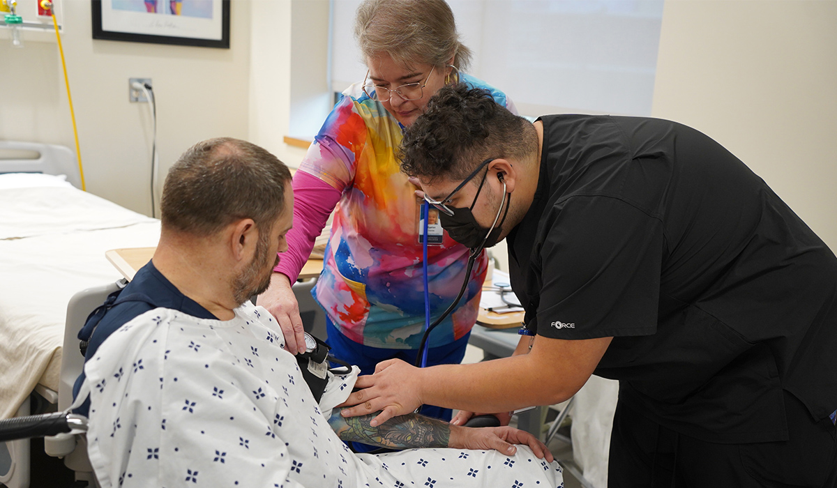 A nursing assistant student checks the pulse of a patient who is seated in a wheelchair.
