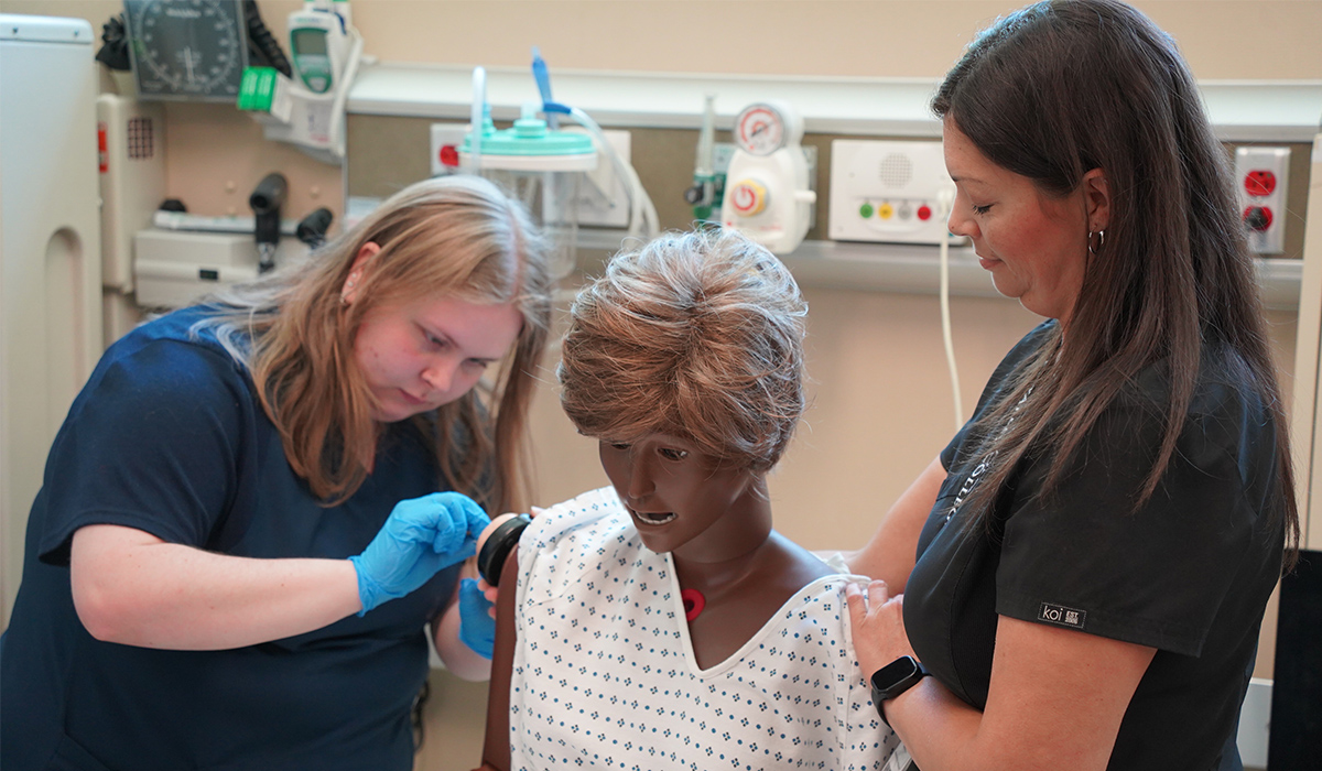 A medical assistant student practices giving a manikin a shot while an instructor holds the manikin upright.