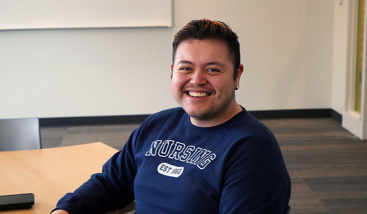 A student is seated at a table in a classroom and wearing a blue sweatshirt with "Nursing" printed across the front.