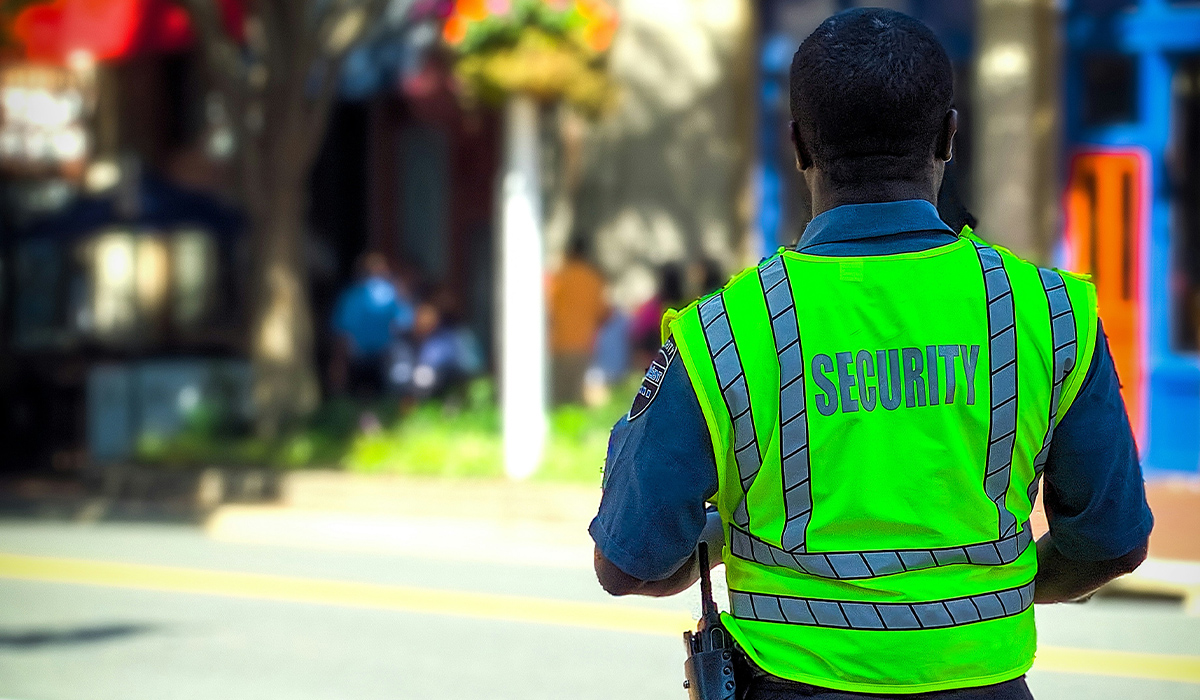 A view from behind a security officer who is wearing a bright green security vest and standing on a sunny street.
