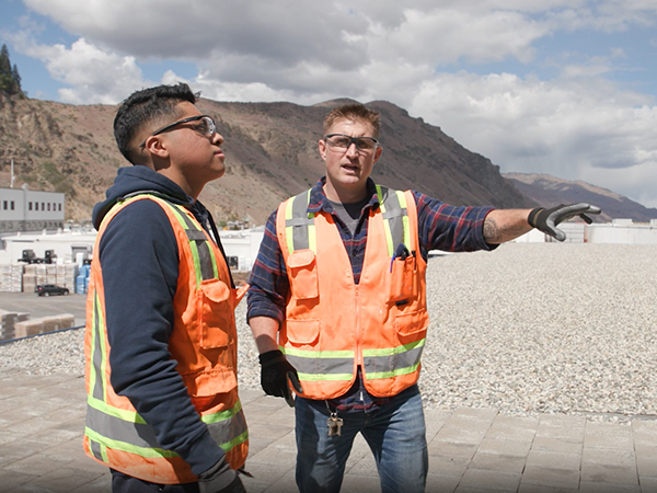 Two refrigeration technicians wearing bright orange vests, safety glasses, and gloves stand outside conversing.