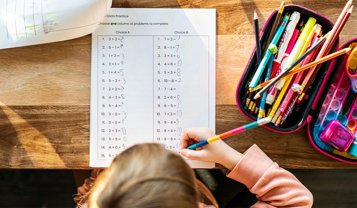 An overhead view of a child sitting at a desk with a math worksheet in front of her.