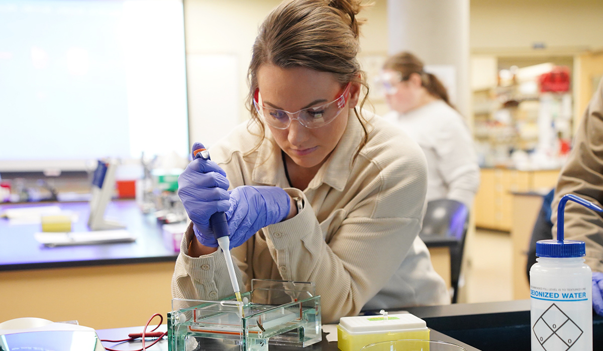 A student wearing a white lab coat and safety glasses conducts an experiment in a biology lab.