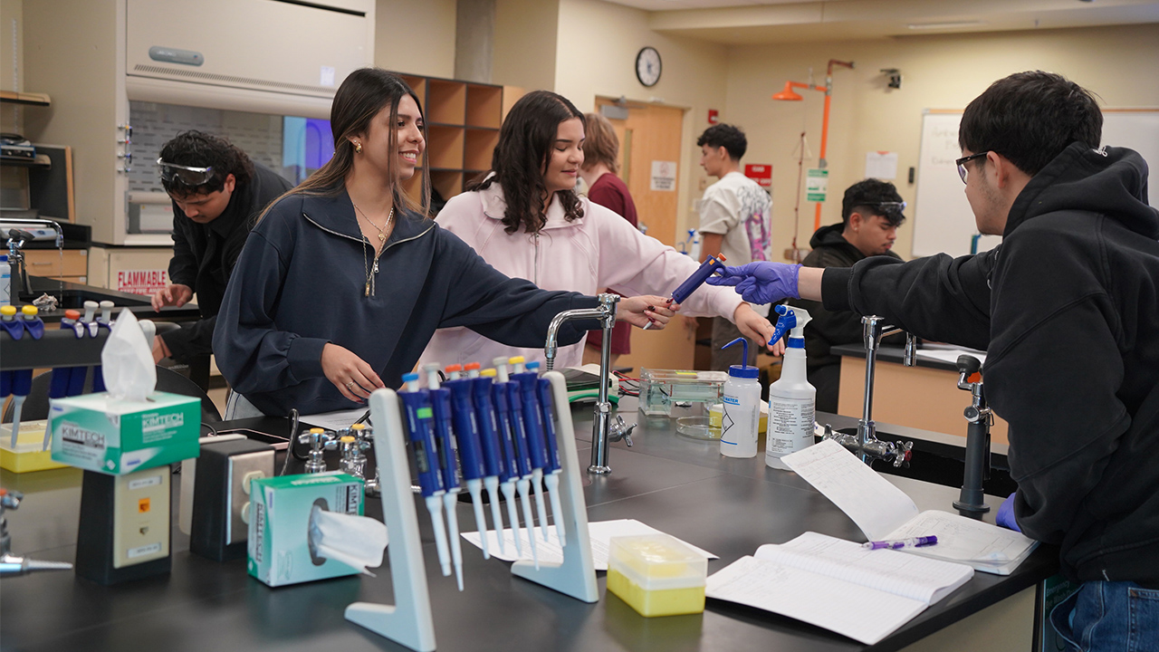 One student passes another student a science tool in  a science lab.