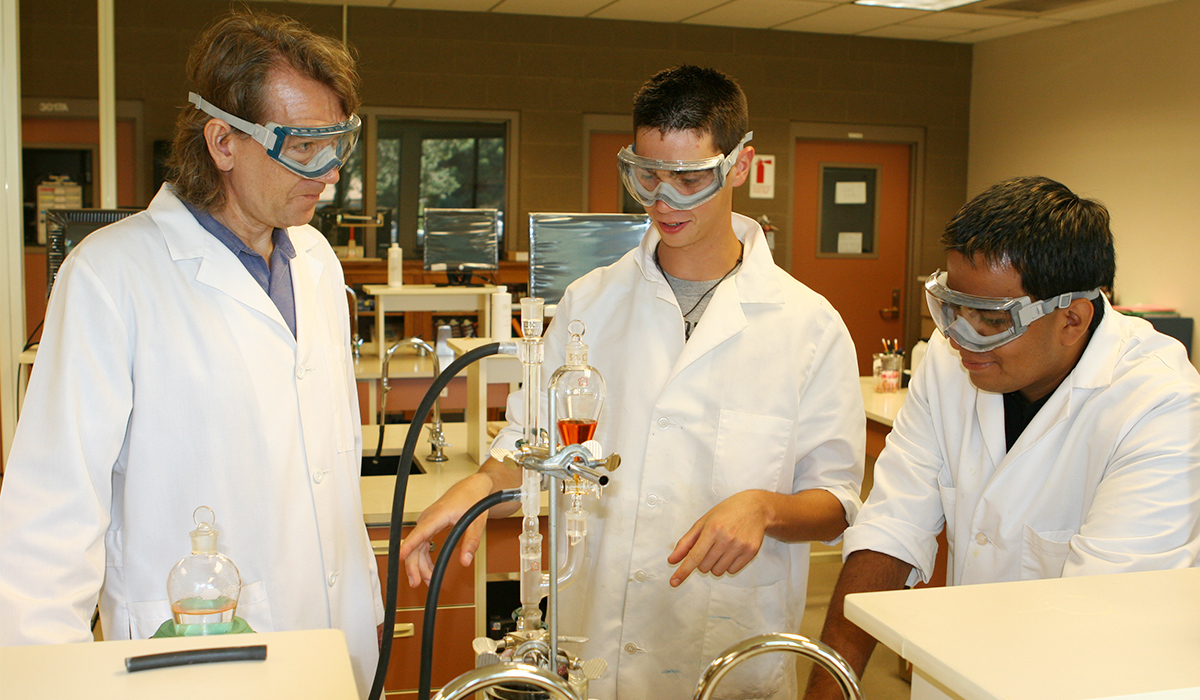 A WVC science professor wearing a white lab coat and safety glasses observes two students conducting a chemistry experiment.