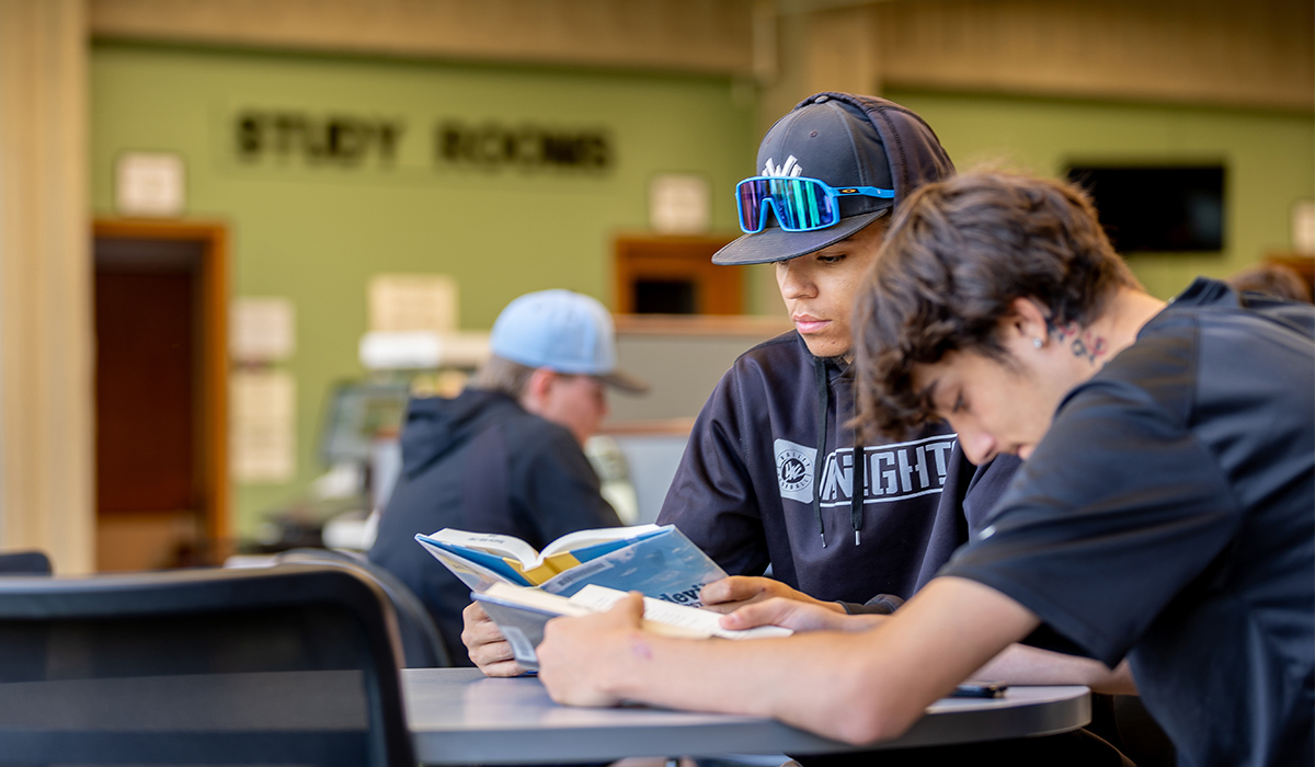 Two students sit at a table in a library and read books.
