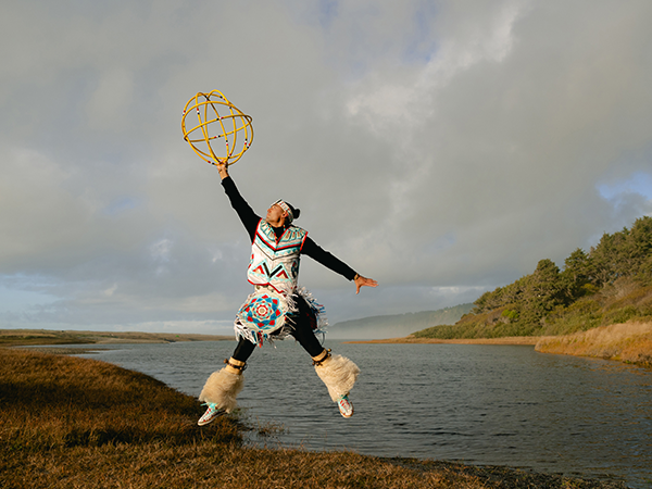 Hoop dancer Eric Hernandez, wearing traditional regalia, is next to a river and leaps into the air holding several hoops.