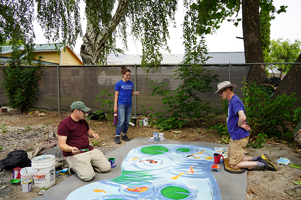 Ellen Bruex stands next to a pond mural in progress with two mural class students.