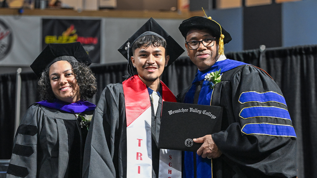 A TRIO graduate wearing a red and white TRIO stole stands with the college president and a trustee member on the stage at the commencement ceremony.