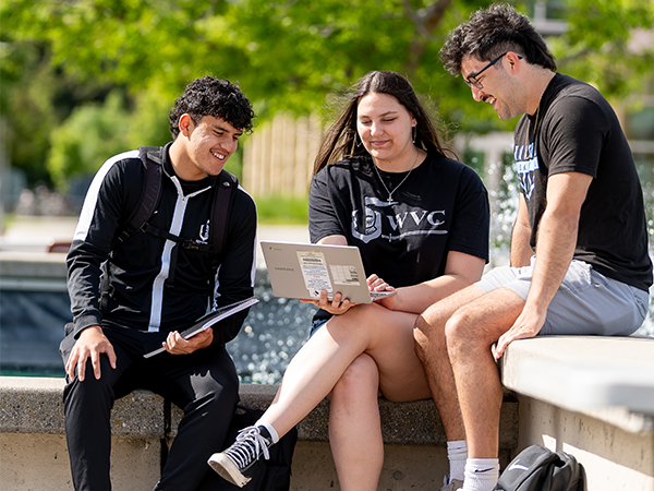 Three WVC students wearing black and white Knights athletic gear sit by the campus fountain and study a laptop.